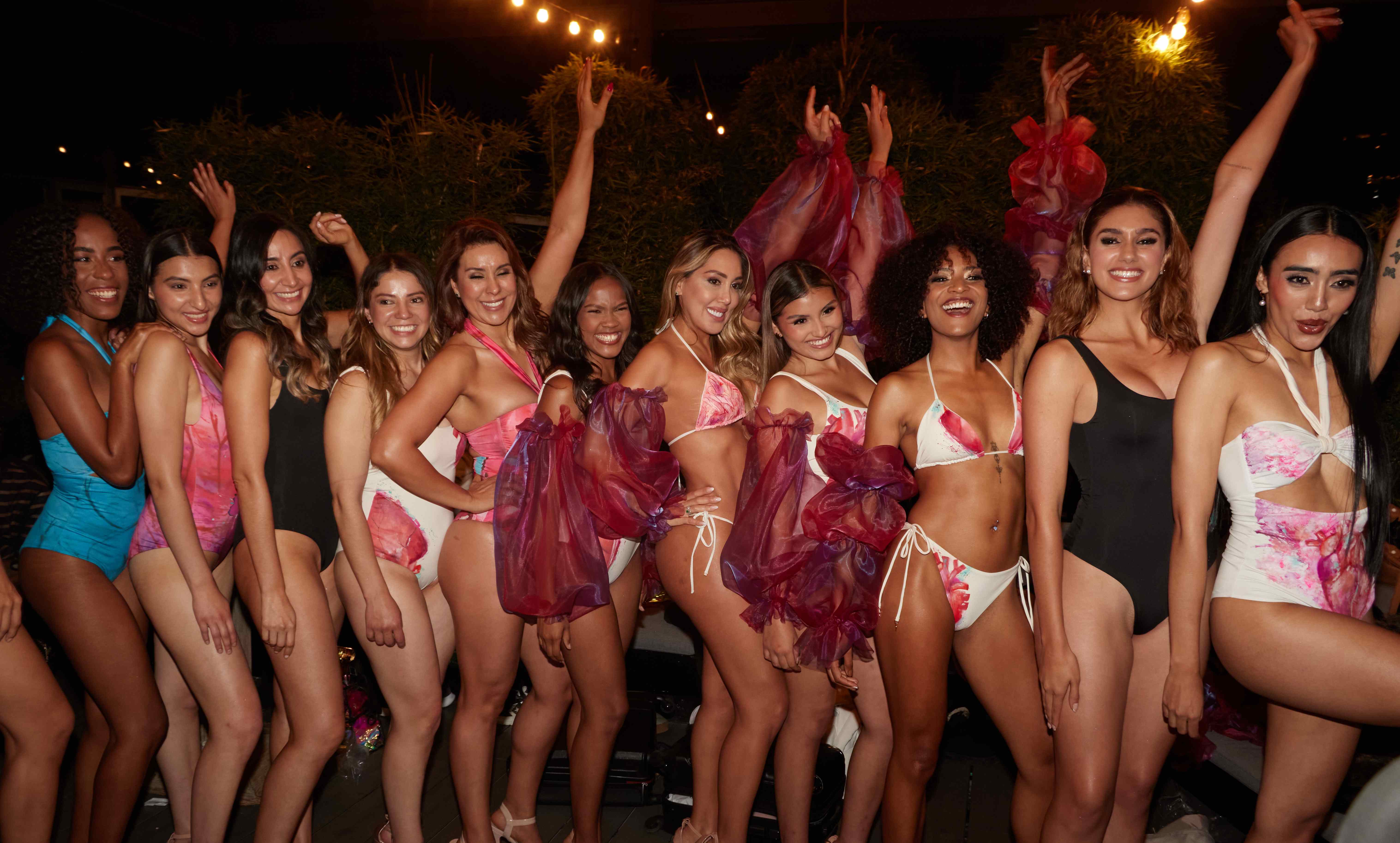 A large group of women of different bodies and skin tones smiling together, arms raised, at an evening rooftop gathering